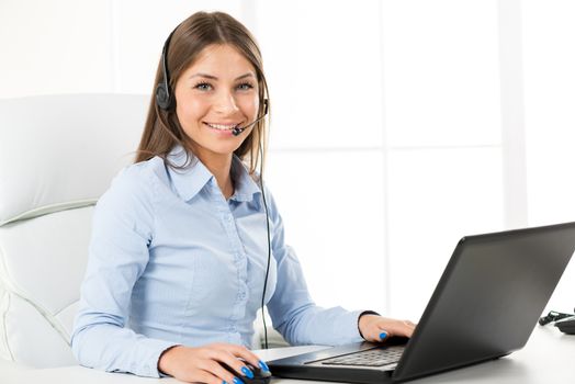 Young businesswoman with headset typing on a laptop in the office with a smile on her face looking at the camera.