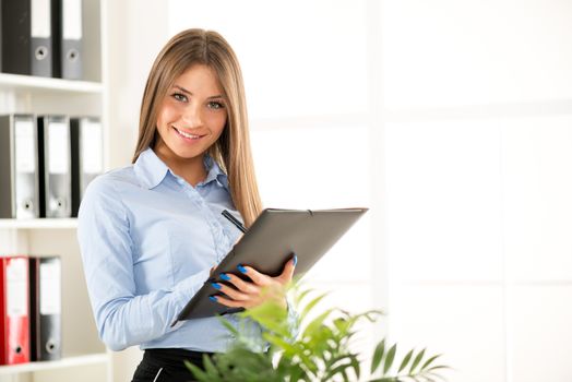 Young smiling businesswoman standing in an office and write on the folder that holds in her hands, in the background you can see the shelves with binders.