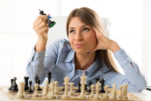 Young pretty businesswoman sitting at the table over a chessboard resting on her hand looking at the black king chess piece that holds in the other hand.