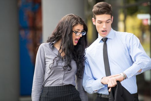 Couple of young business people, elegantly dressed, attractive businesswoman with glasses and a businessman, with an expression of astonishment on their faces, looking at businessman wristwatch.