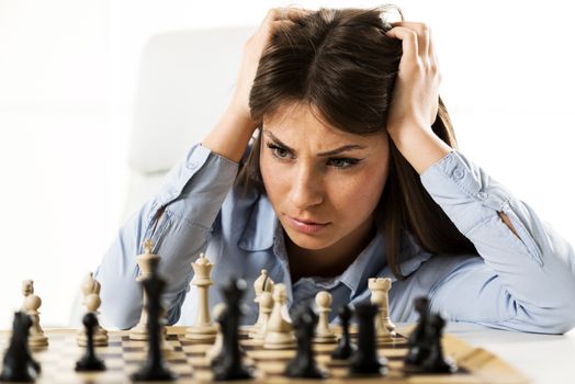 Young worried businesswoman in front of a chess table, holding her hands to her head looking at the chessmen worrying about the validity of her moves.