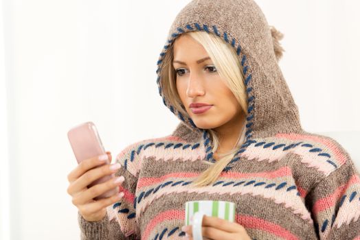 Beautiful young woman in a wool sweater with a hood on her head, looking at mobile phone that holds in her hand, and in the other hand holding a cup.