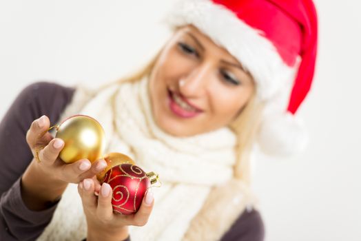 Cute blonde girl, out of focus, with a cap of Santa Claus and a warm scarf around her neck, holding a decorative balls that are in the foreground and with a smile on her face looking at them.