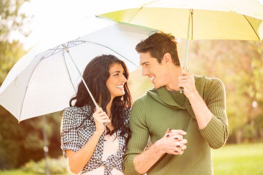 Young heterosexual couple walking in park holding hands and carrying umbrellas.