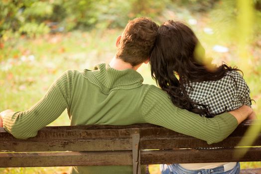 Photo of a young couple, their backs turned to the camera, sitting on a bench, leaning their heads on each other.