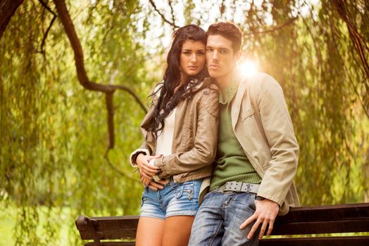 Young attractive heterosexual couple standing leaning on a bench in the park looking at the camera while the sun shines from behind.