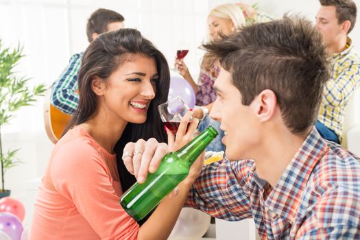 A young girl and guy at home party, drinking with crossed arms, smiling looking at each other, in the background you can see their friends who are sitting on the couch.
