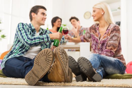 Young couple at house party with faces out of focus in the foreground are the soles of their shoes, sit on the floor, knocking the toasting glasses, in the background you can see another couple.
