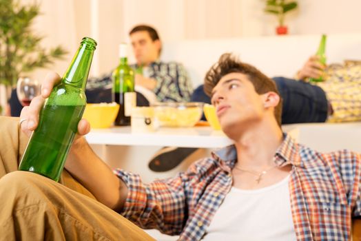 A young man with a face out of focus, tired of the party sitting on the floor leaning against the table, holding a bottle of beer, which is in the foreground. In the background you can see two hungover guy lying on the couch.