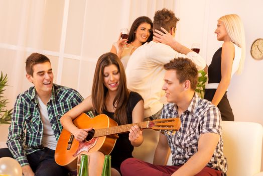 Girl playing acoustic guitar at house party, sit on the couch between two guys, and behind them is a guy who speak with two girls.