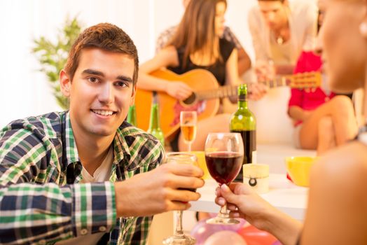 A young man with a glass of wine on the house party toast with a girl, and in the background you can see a group of young people gathered around the girl with an acoustic guitar.