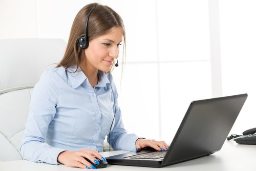 Young businesswoman with headset, smiling, typing on a laptop in the office.