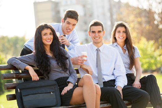 Small group of business people, elegantly dressed, on a coffee break in the park, sitting on a bench, enjoying the beautiful weather and with a smile on their faces looking at the camera.