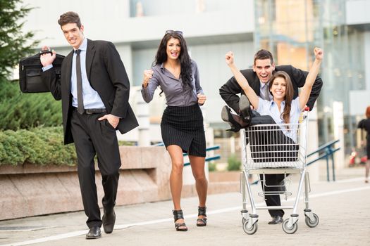 Group of young business people on the street near a supermarket in a hurry to sell, pushing their colleague in a shopping trolley.