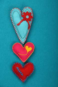 Felt craft and art, three handmade stitched toy hearts, pink, red and teal on blue felt background