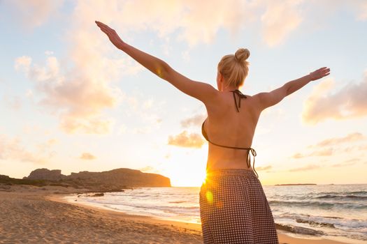 Relaxed woman, arms rised, enjoying sun, freedom and life an beautiful beach in sunset. Young lady feeling free, relaxed and happy. Concept of vacations, freedom, happiness, enjoyment and well being.