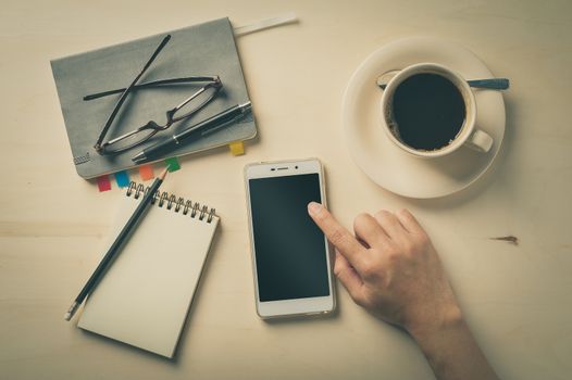 Male finger touching on smartphone screen with notebooks, pen, and coffee cup beside on wood table in morning time with vintage filter effect