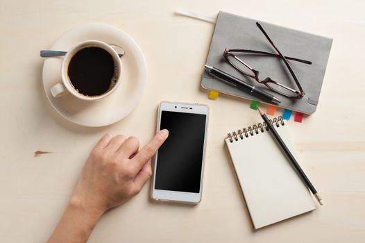 Male finger touching on smartphone screen with notebooks, pen, and coffee cup beside on wood table in morning time