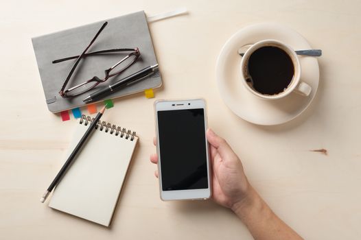 Male right hand holding smartphone with notebooks, pen, and coffee cup beside on wood table in morning time