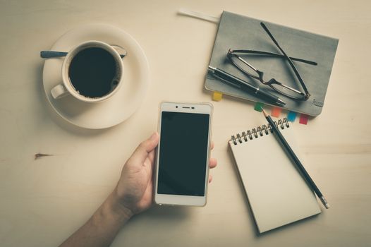 Male left hand holding smartphone with notebooks, pen, and coffee cup beside on wood table in morning time with vintage filter effect