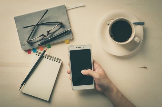 Male finger touching on smartphone screen with notebooks, pen, and coffee cup beside on wood table in morning time with vintage filter effect