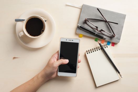 Male finger touching on smartphone screen with notebooks, pen, and coffee cup beside on wood table in morning time