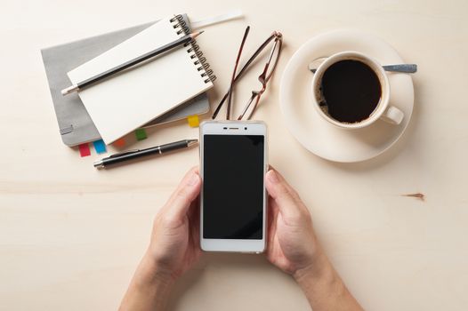 Male hand holding smartphone with notebooks, pen, and coffee cup beside on wood table in morning time