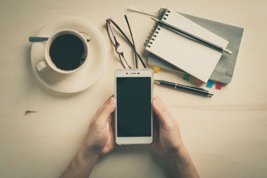 Male hand holding smartphone with notebooks, pen, and coffee cup beside on wood table in morning time with vintage filter effect