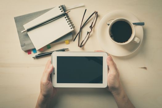 Male hand holding small tablet pc with notebooks, pen, and coffee cup beside on wood table in morning time with vintage filter effect