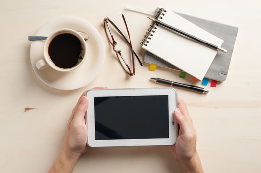 Male hand holding small tablet pc with notebooks, pen, and coffee cup beside on wood table in morning time