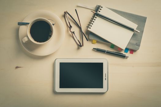 Small tablet pc, notebooks, pen, glasses, and coffee cup on wood table in cafe at morning time with vintage filter effect