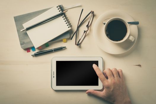 Male hand touching on small tablet pc screen with notebooks, pen, and coffee cup beside on wood table in morning time with vintage filter effect