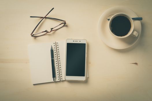 Opened notebook with blank area, pen, smart phone, and coffee cup on wood table in morning time with vintage filter effect