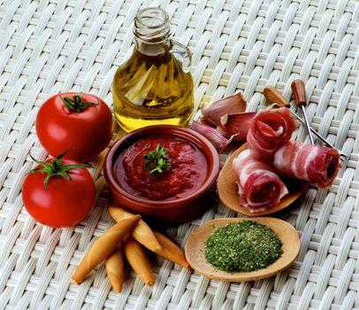 Tapas and Ingredients with Smoked Bacon, Tomatoes and Tomato Sauce, Bread Sticks, Spices and Olive Oil closeup on Wicker background