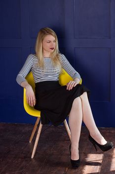 Blond girl in a long skirt and a vest sits on a chair next to the blue walls