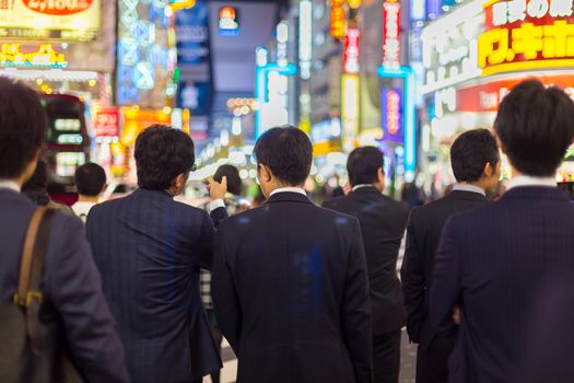 Japanese corporative business people in suits, waiting in rush hour on crossroad in Shinjuku business district, Tokyo, Japan. Blured advertising boards illuminated in the background.