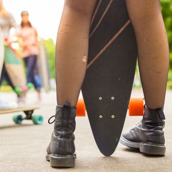 Teenagers practicing long board riding outdoors in skateboarding park. Detail of  legs of girl wearing black boots and stockings holding long board in foreground. Active urban life. Urban subculture.