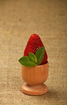 One big red mellow strawberry and green fresh mint leaves in wooden eggcup holder on natural burlap jute canvas background