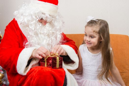 Santa Claus helps to untie the ribbon gift, little girl happily looking at him