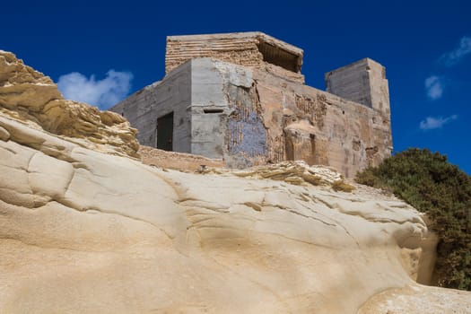 Sand color of the rocks with curved shape and an old building. Coast of island Malta, city Marsaskala. Blue sky with several white clouds.
