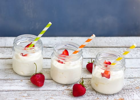 Three jars of yogurt with strawberries and colored tubes on a wooden board