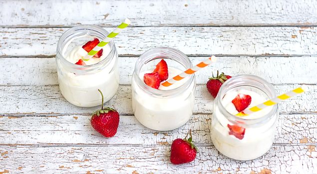 Three jars of yogurt with strawberries and colored tubes on a wooden board