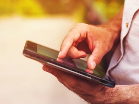 Smart casual man with digital tablet outdoors, close up. Focus on finger. Sunkissed effect with sunflares. Modern remote work concept.