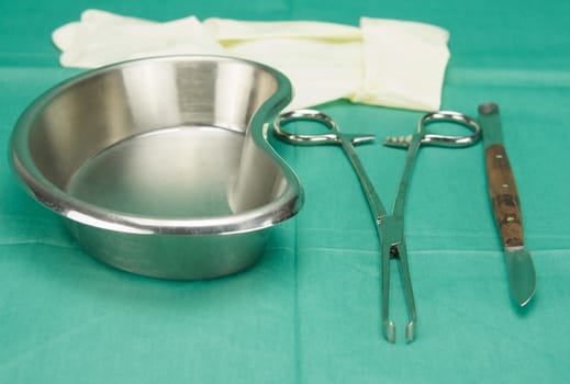 Surgical clamp and knife placed near kidney shape bowl on green fabric.