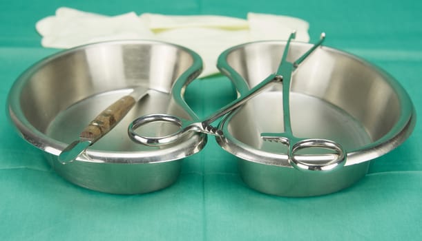 Surgical clamp and knife placed on stainless kidney shape bowl on green fabric.