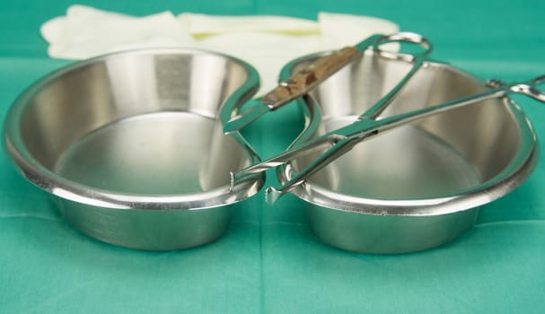 Surgical clamp and knife placed on stainless kidney shape bowl on green fabric.