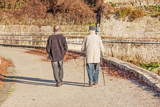 Two older people walking - one with walking sticks. View from behind.