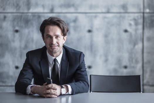 Portrait of a businessman with smartphone sitting behind the table over concrete wall background