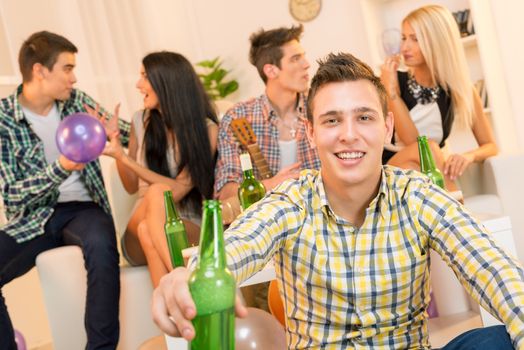 A young guy at a home party, with a smile on his face looking at the camera sitting on the floor holding a bottle of beer with which toasts the viewer. In the background you can see his friends sitting on the couch.