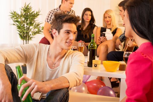 A group of young people in house party. In the first plan is handsome young man sitting on the floor, talking to a girl, and in the background a group of young people sitting on the couch.